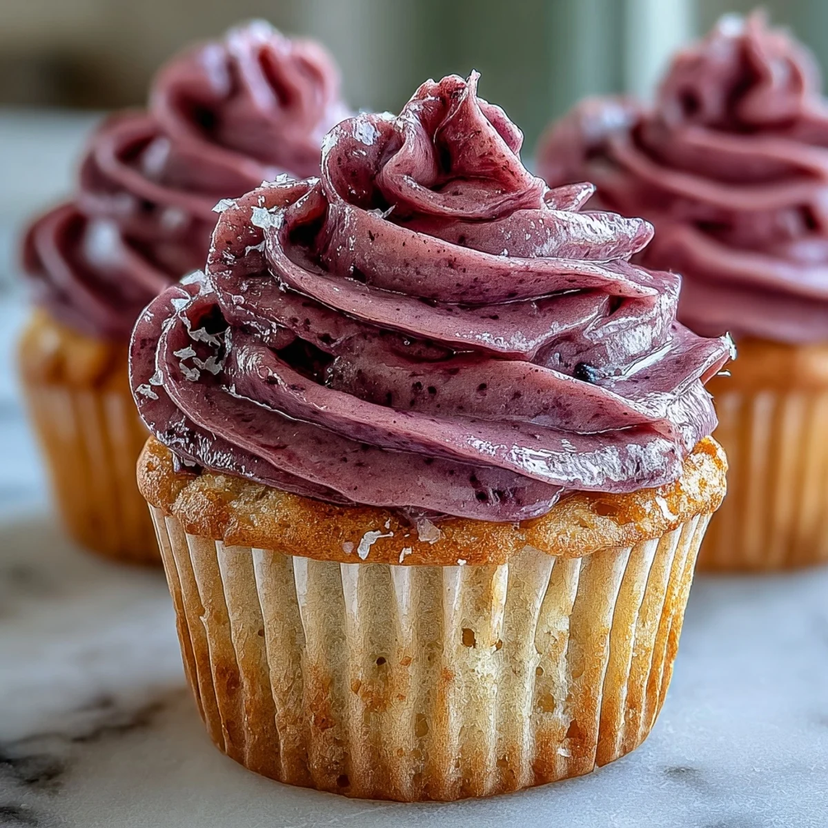 Moist olive oil cupcakes topped with swirls of purple black currant frosting and lemon zest, served on a ceramic plate.