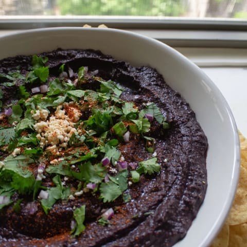 Thick, vibrant spicy black bean dip surrounded by crunchy tortilla chips and colorful vegetable sticks, perfect for party snacking or game day.